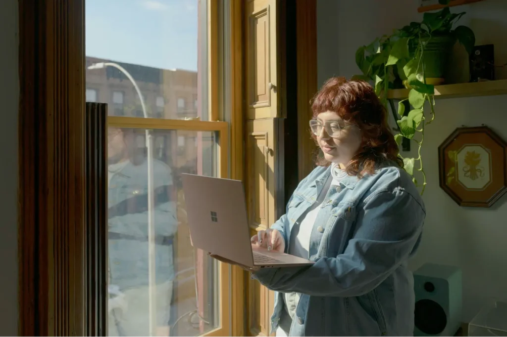 A woman standing by a window, holding a laptop open having a virtual meeting.